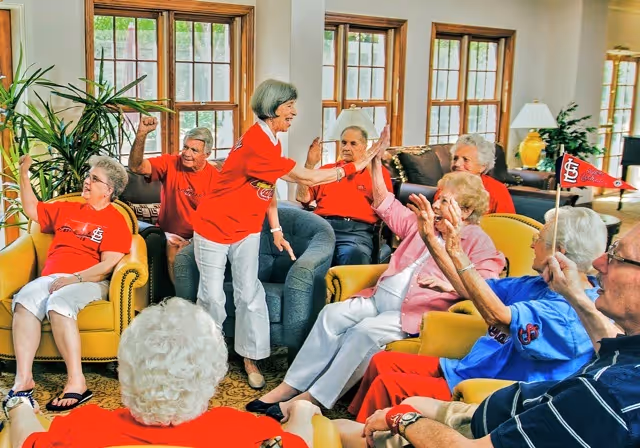 A group of elderly people sitting and standing in a bright living room with large windows, engaging in a lively activity. Some are wearing red shirts with a sports logo, and one person is holding a small flag. They appear to be cheering and interacting happily with each other.