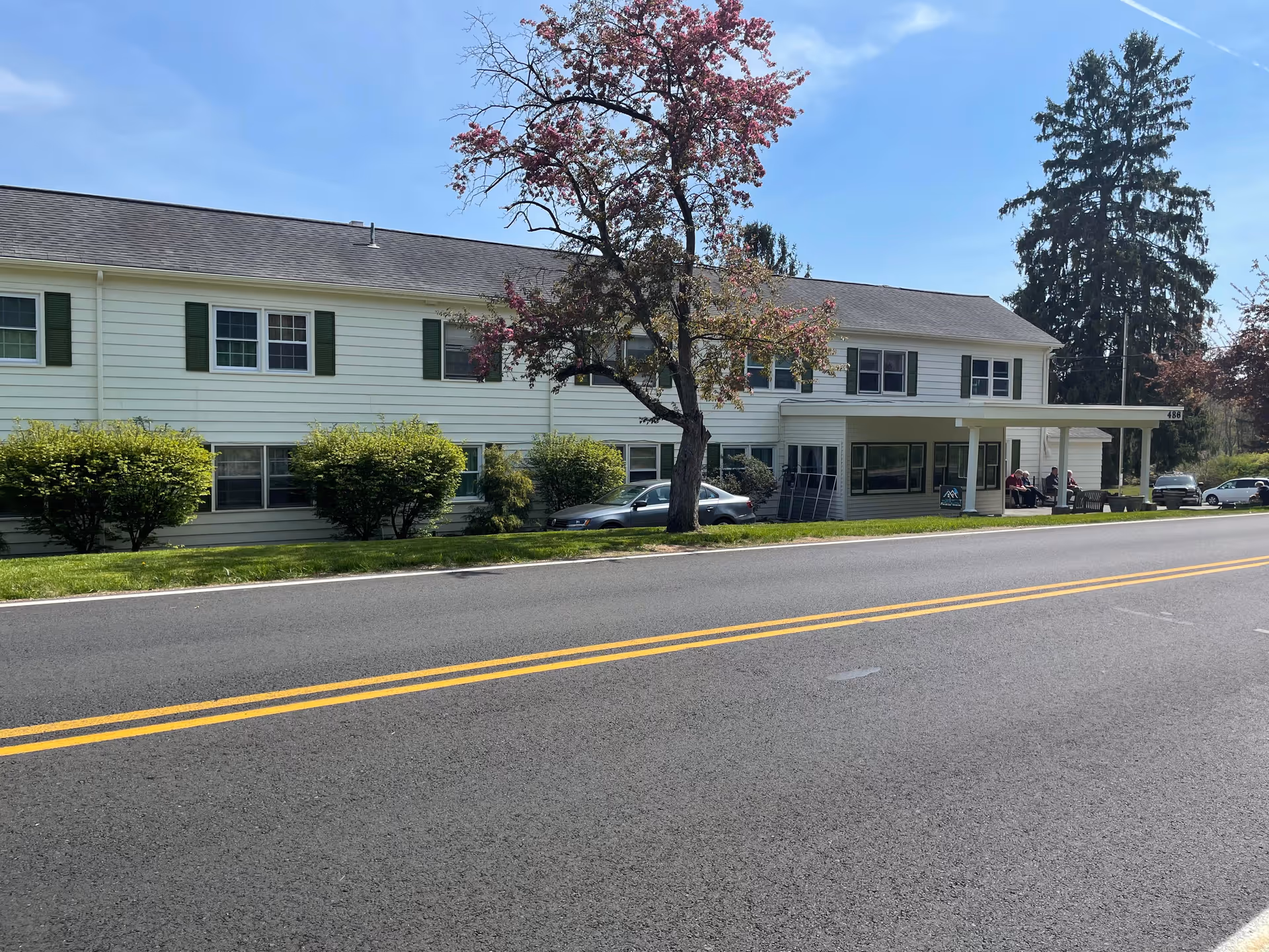 Exterior view of a two-story residential care facility building with white siding and green shutters, a tree with pink blossoms in front, several bushes, parked cars, and a covered entrance with people sitting nearby under a clear blue sky.