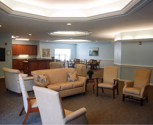 Spacious senior living common room with sofas, armchairs, dining tables and a kitchenette area under recessed ceiling lights.