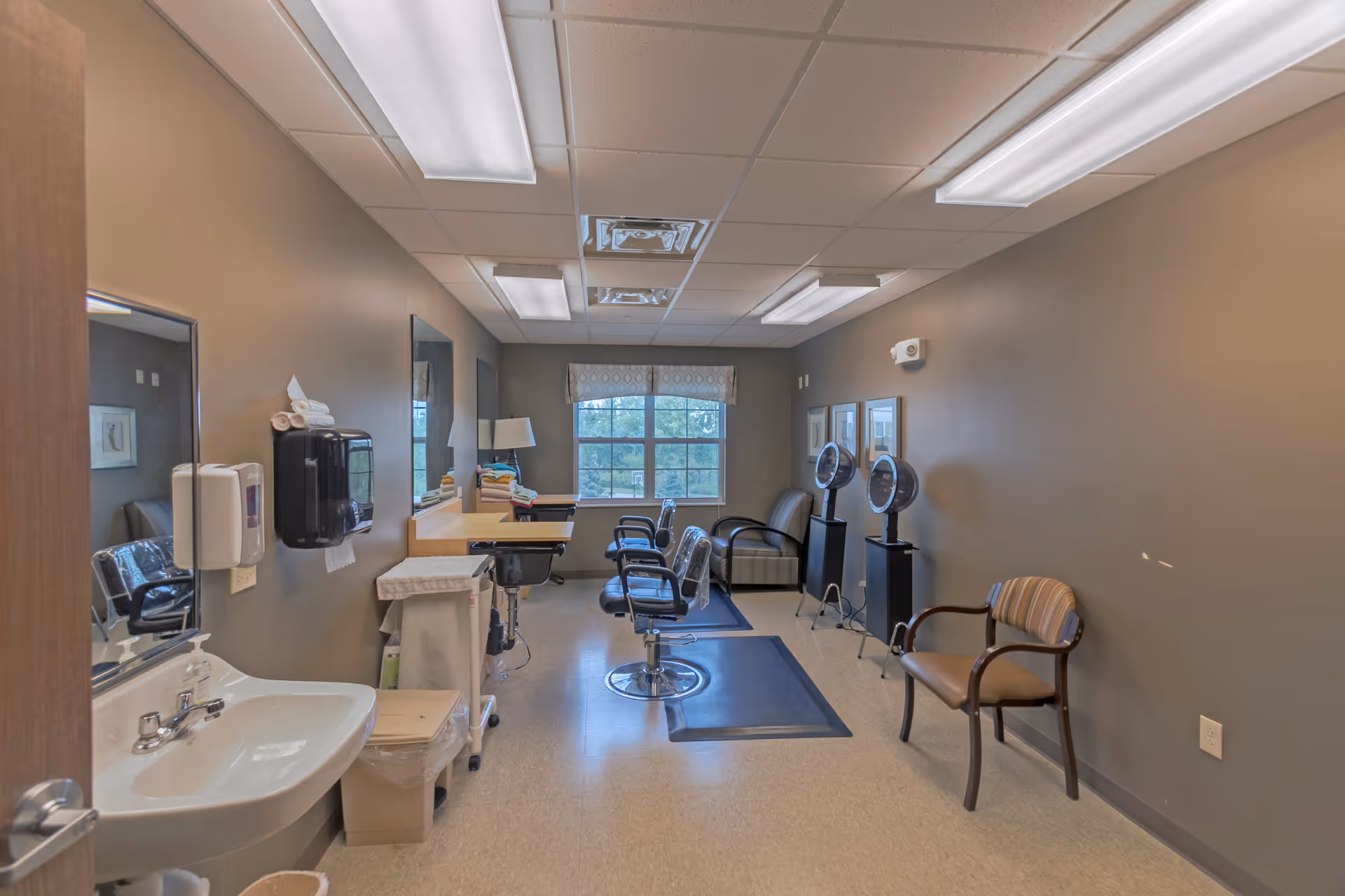 Interior view of a hair salon area in a senior living facility with salon chairs, hair dryers, a sink, mirrors, and a window with a valance. The room has beige walls and ceiling lights.