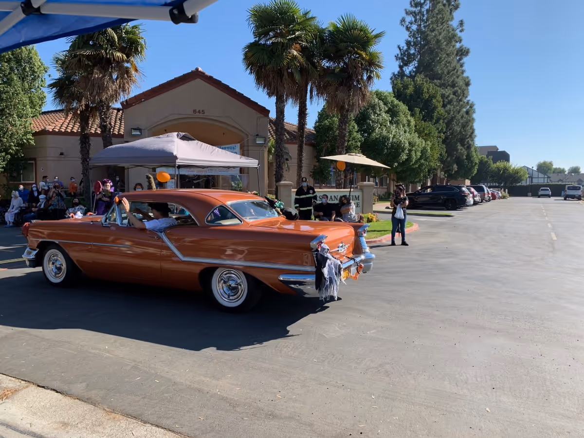 A vintage orange car decorated with Halloween-themed items is driving in front of a building with palm trees and people gathered under a canopy and around the parking lot on a sunny day.