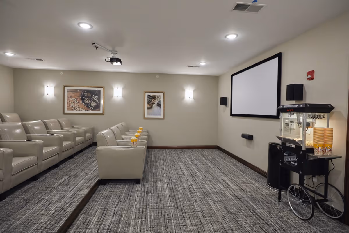 A senior living facility movie theater room with two rows of beige leather recliner chairs, each with a popcorn container on the armrest. The room has a large projector screen on the wall, a popcorn machine with cups on a cart, framed pictures on the walls, and ceiling lights.
