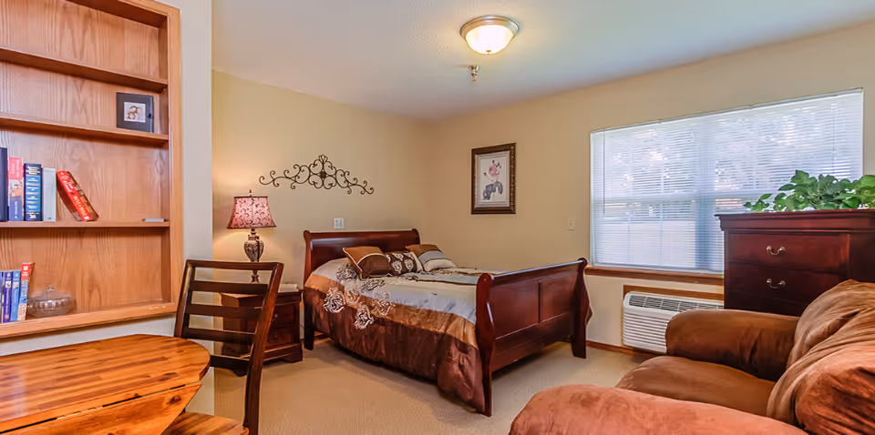 A cozy senior living bedroom featuring a wooden bed with brown and beige bedding, a wooden nightstand with a decorative lamp, a wooden bookshelf with books, a wooden table with a chair, a brown upholstered armchair, a wooden dresser with a green plant on top, and a window with blinds letting in natural light.