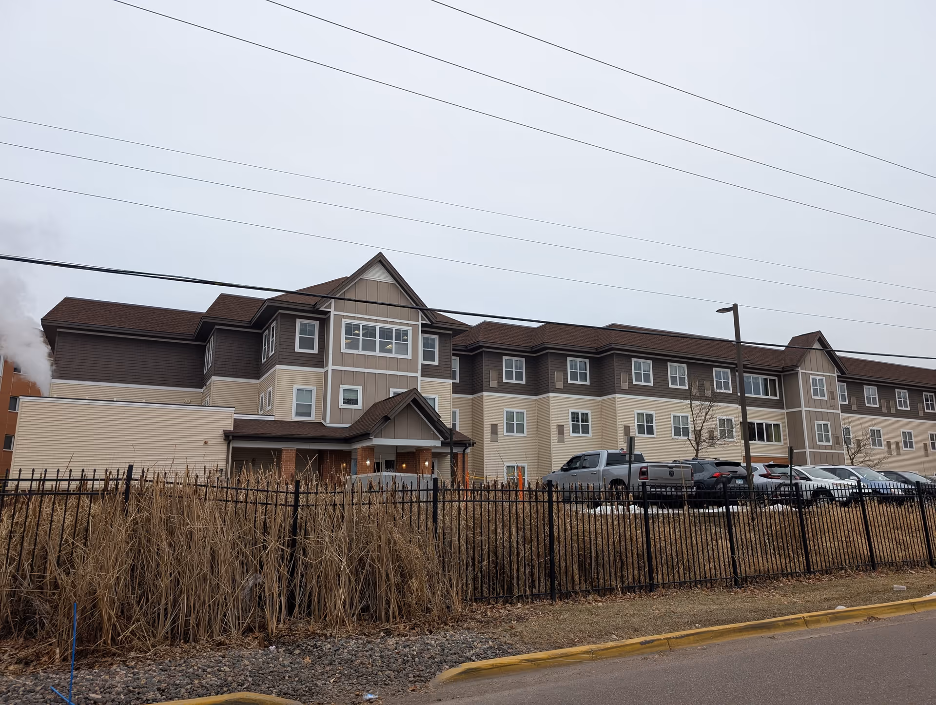 Exterior view of a three-story senior living facility building with beige and brown siding, multiple windows, a small covered entrance, a black metal fence, parked cars, and dry tall grass in front under an overcast sky.