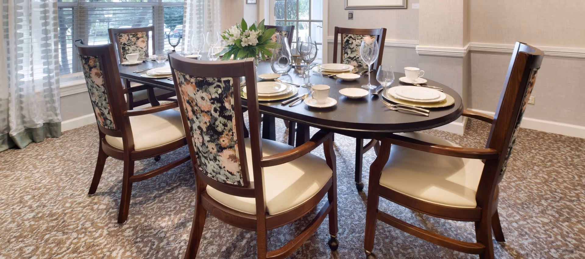 A dining room with a dark wooden oval table set for six people. The table is arranged with white plates, cups, wine glasses, and silverware. There is a floral centerpiece in the middle of the table. The chairs have wooden frames with floral-patterned backs and cream-colored cushions. The room has carpeted flooring and large windows with sheer curtains allowing natural light to enter.