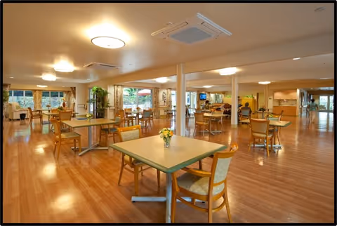 A spacious dining area with multiple tables and chairs arranged on a polished wooden floor. The room is well-lit with ceiling lights and has large windows allowing natural light to enter. There are plants and decorative elements near the windows, and a few people are seated at the tables in the background.
