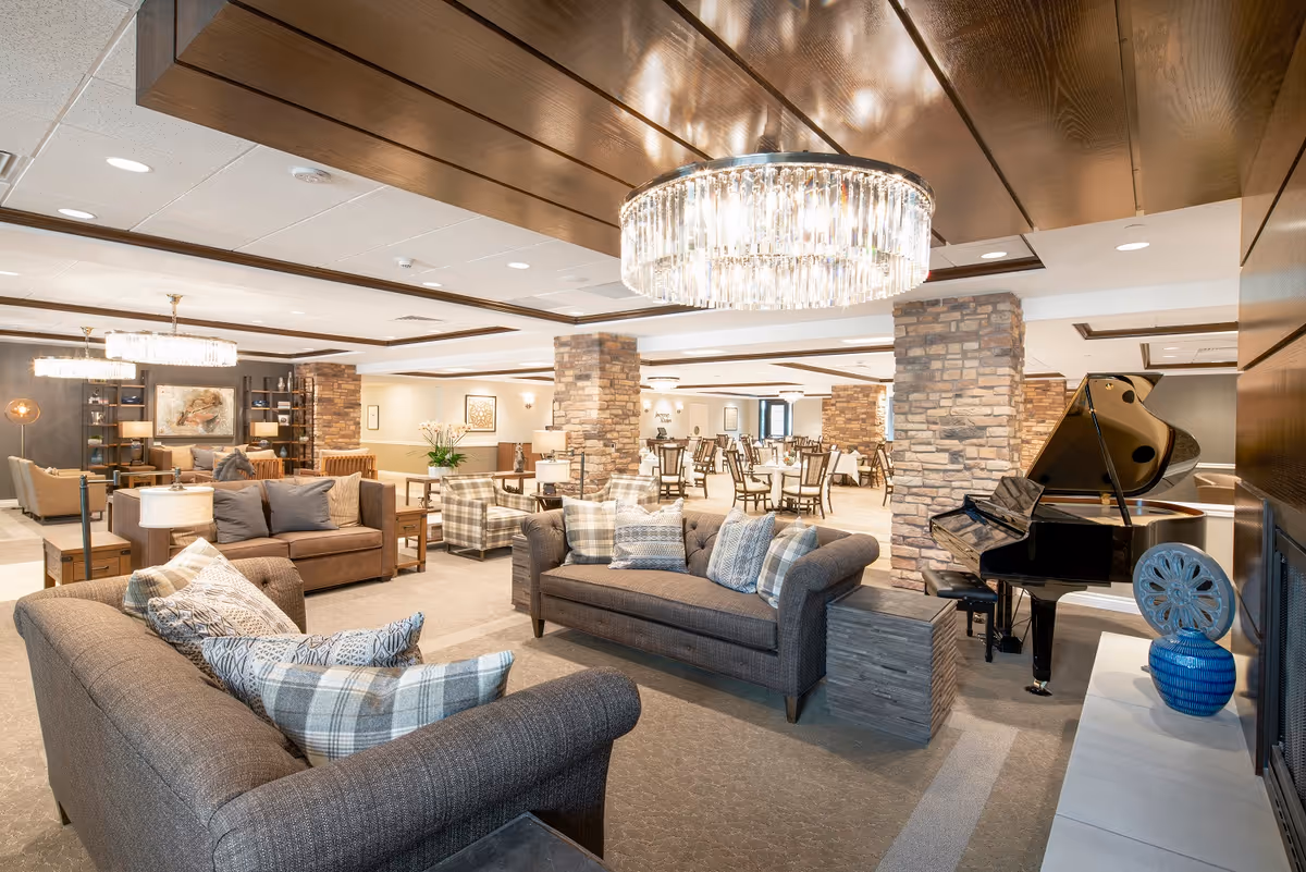 A spacious and well-lit senior living facility common area featuring comfortable sofas with patterned pillows, armchairs, a grand piano, and a dining area with multiple tables and chairs. The room has stone pillars, wooden ceiling accents, and elegant chandeliers providing warm lighting.