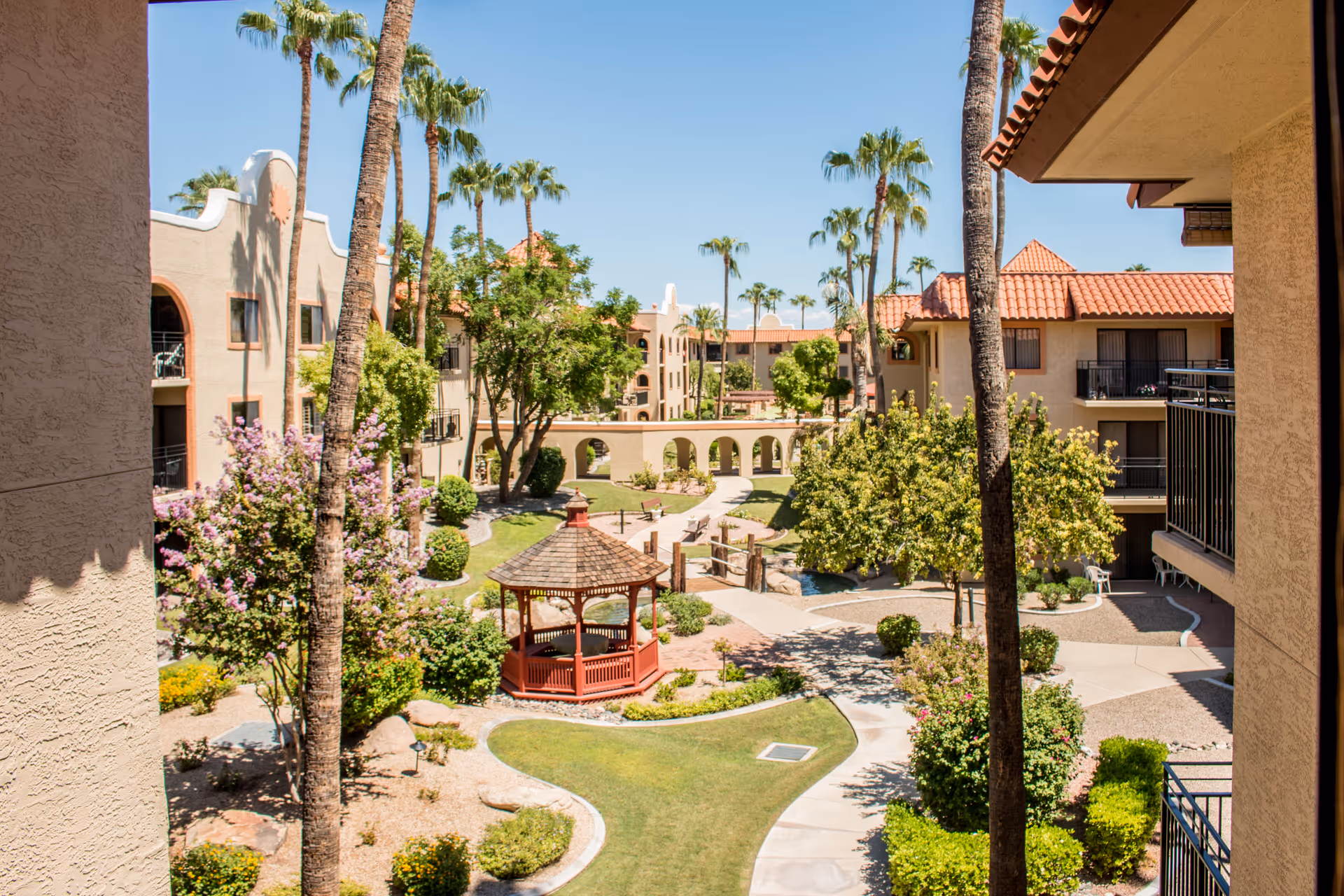 View of a landscaped courtyard in a senior living facility with palm trees, flowering bushes, a red gazebo, walking paths, and beige buildings with red tile roofs under a clear blue sky.