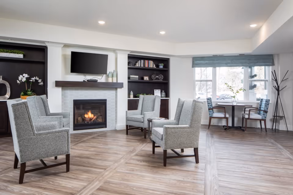 A bright and modern living room with four gray upholstered armchairs arranged around a lit fireplace. Above the fireplace is a mounted flat-screen TV. Built-in dark wood shelves flank the fireplace, decorated with books and plants. A small table with two chairs is positioned near a large window with a blue valance, allowing natural light to fill the room. The floor is wood with a light finish.