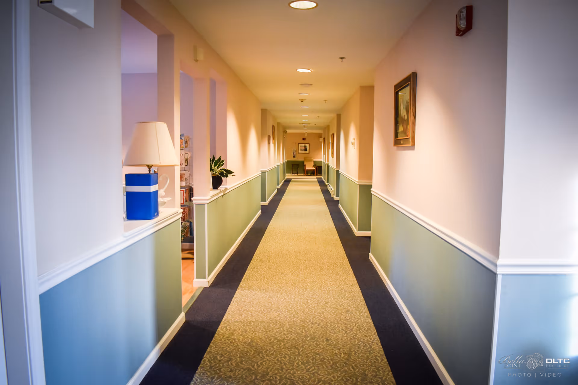A long, well-lit hallway in a senior living facility with light pink upper walls and green lower walls separated by white trim. The carpet has a patterned beige center with dark blue borders. There are framed pictures on the walls, a lamp on a ledge to the left, and a plant further down the hallway. At the far end, there are chairs and a table.