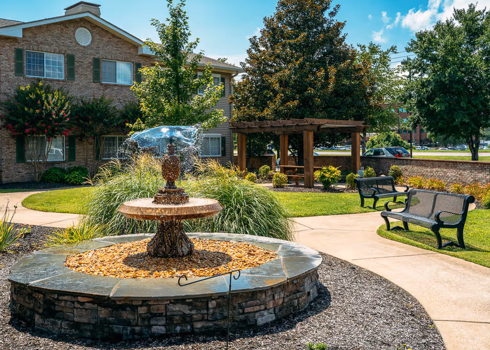 Outdoor garden area at The Bridge at Columbia featuring a stone fountain with water flowing, surrounded by landscaping with green grass, bushes, and trees. There are two black metal benches along a curved concrete pathway and a wooden pergola with picnic tables in the background. A brick building with green shutters is visible behind the garden.