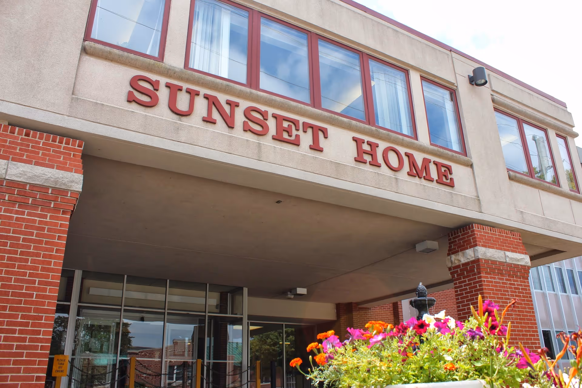 Exterior entrance of a building with a 'SUNSET HOME' sign above the portico and flowers in the foreground.