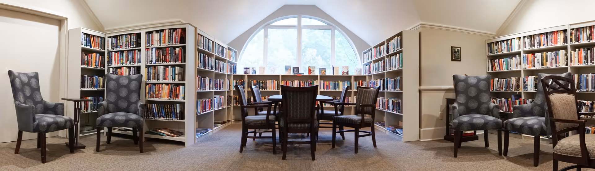A cozy library room with bookshelves lining the walls filled with books. There are several comfortable chairs arranged in pairs on either side of the room, and a round table with six chairs in the center near a large arched window letting in natural light.