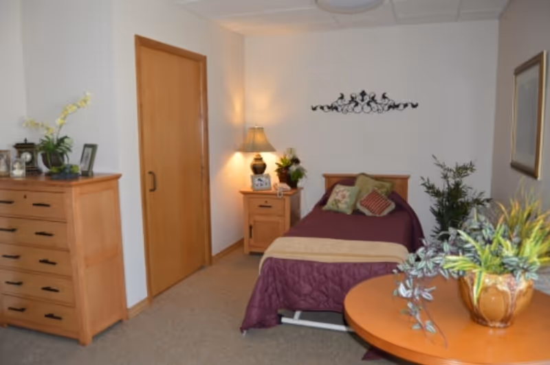 A cozy bedroom with a single bed covered in a purple quilt and decorative pillows. There is a wooden nightstand with a lamp, a small plant, and a photo frame next to the bed. A wooden dresser with plants and framed photos is against the wall near a closed wooden door. A round wooden table with a potted plant is in the foreground, and a framed picture hangs on the wall.