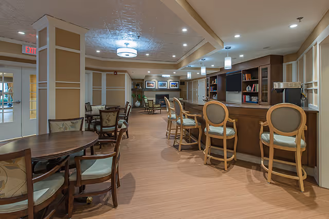 Interior view of a senior living facility lounge area with a wooden bar counter and four cushioned bar stools. There are several round tables with chairs arranged around the room. The space is well-lit with ceiling lights and pendant lights above the bar. In the background, there is a seating area with armchairs and framed artwork on the wall.