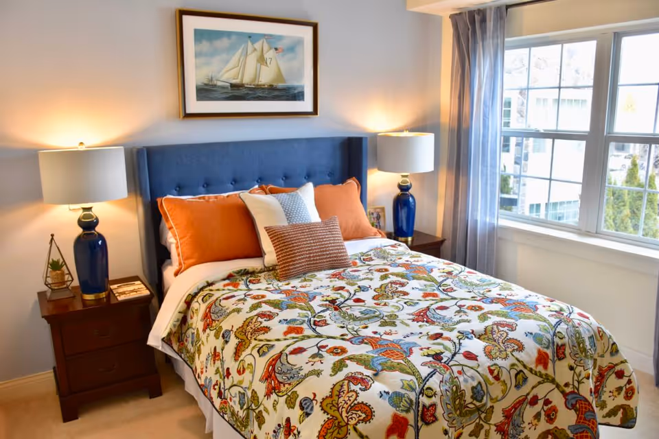 A cozy bedroom with a blue upholstered headboard, a bed adorned with a colorful floral patterned quilt and multiple pillows in orange, white, and brown. There are two wooden nightstands on either side of the bed, each with a blue lamp and other small decorative items. A large window with sheer curtains lets in natural light, and a framed painting of a sailboat hangs above the bed.