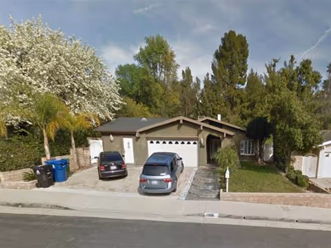 Single-story house with a two-car driveway, trees, and parked cars along the front.