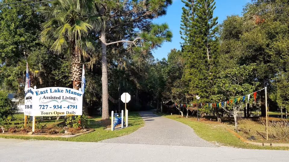 Entrance to East Lake Manor Assisted Living Facility with a large sign displaying the facility name, phone number, and 'Tours Open Daily'. The entrance road is lined with trees and colorful pennant flags on the right side.