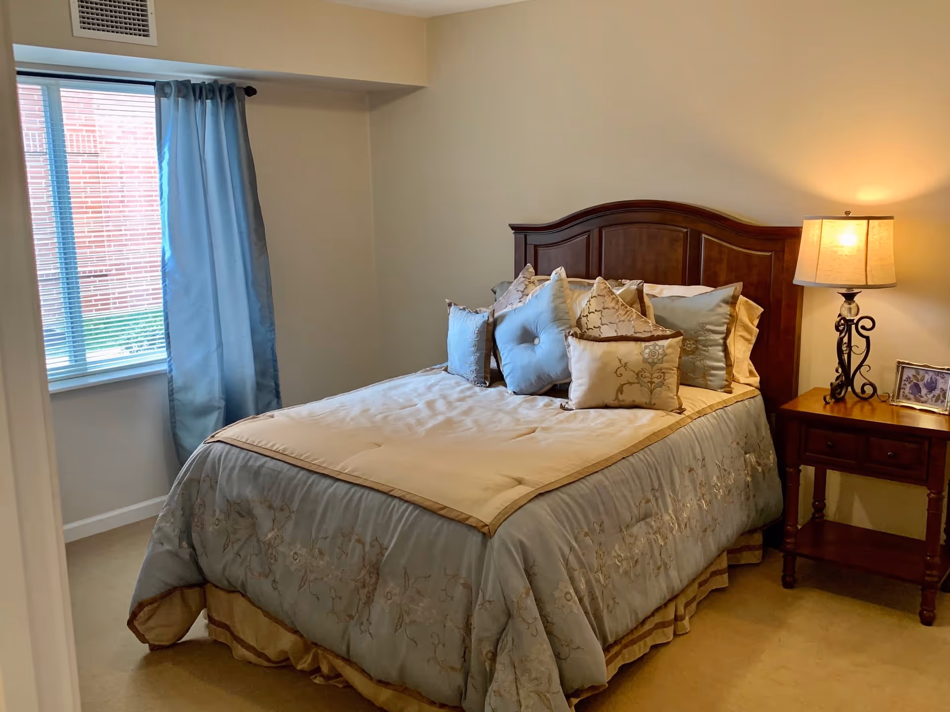 A neatly made bed with a wooden headboard in a bedroom. The bed has a beige and light blue comforter with multiple decorative pillows in matching colors. To the right of the bed is a wooden nightstand with two drawers, a decorative lamp with a beige shade, and a small framed picture. A window with blue curtains is on the left wall, letting in natural light.