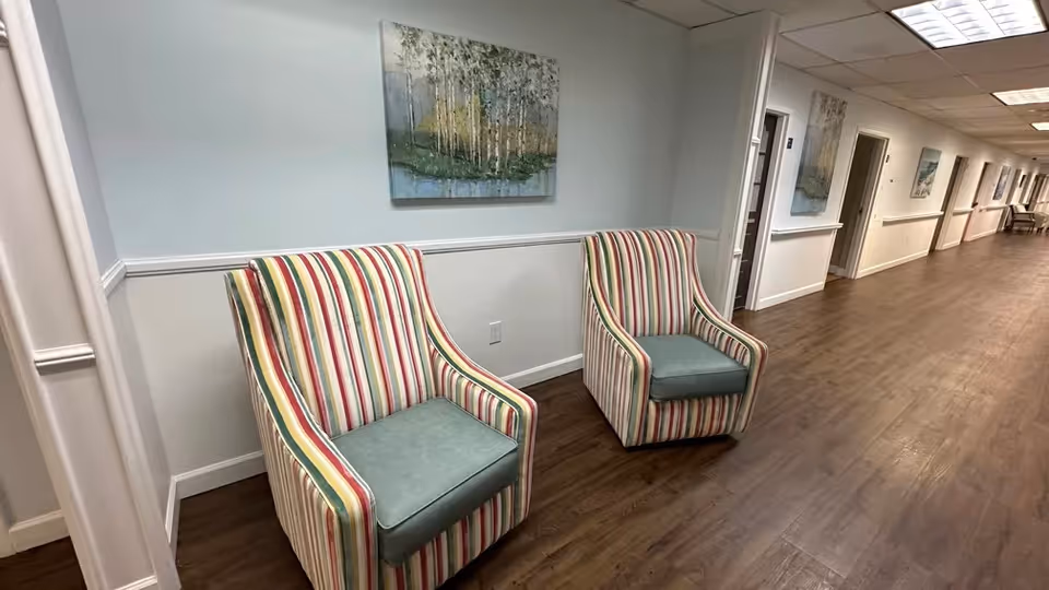 Two striped armchairs sit against a pale blue wall in a long, well-lit hallway with artwork and wood flooring.