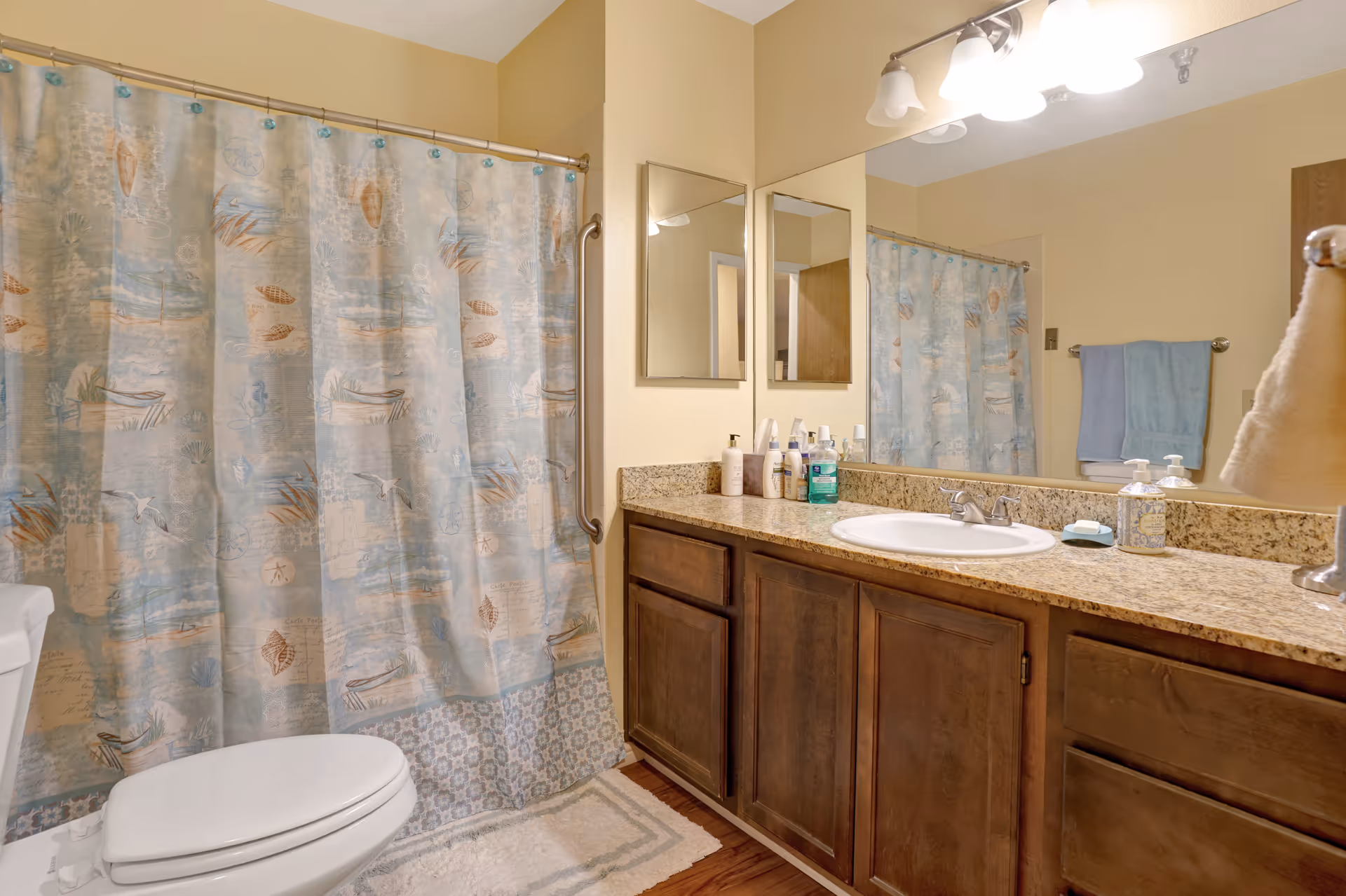 A bathroom with a toilet, a bathtub with a blue and beige patterned shower curtain, a granite countertop with a sink, wooden cabinets underneath, a large mirror above the sink, and various toiletries on the counter. There is a towel hanging on a rack on the wall and a beige wall color.