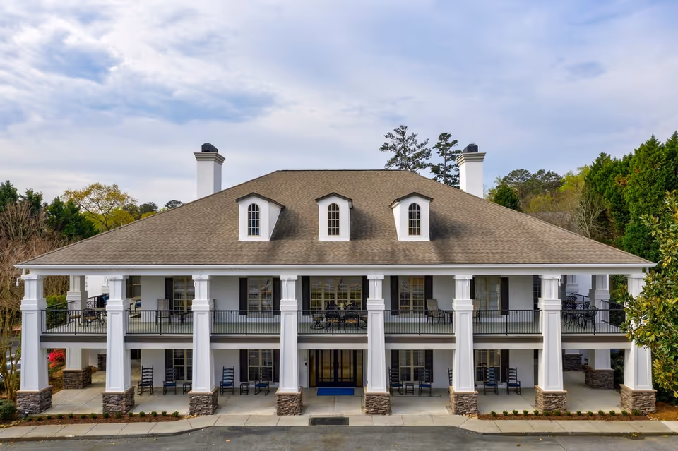 Front exterior of a two-story Colonial-style building with large white columns, a covered veranda with rocking chairs, and a pitched roof with dormer windows.