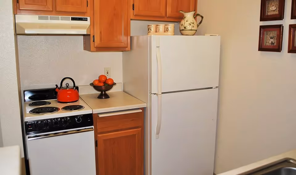 A small kitchen area with wooden cabinets, a white refrigerator, an electric stove with four burners, a red kettle on the stove, a bowl of oranges on the countertop, and decorative items on top of the refrigerator. There are also two framed pictures hanging on the wall.