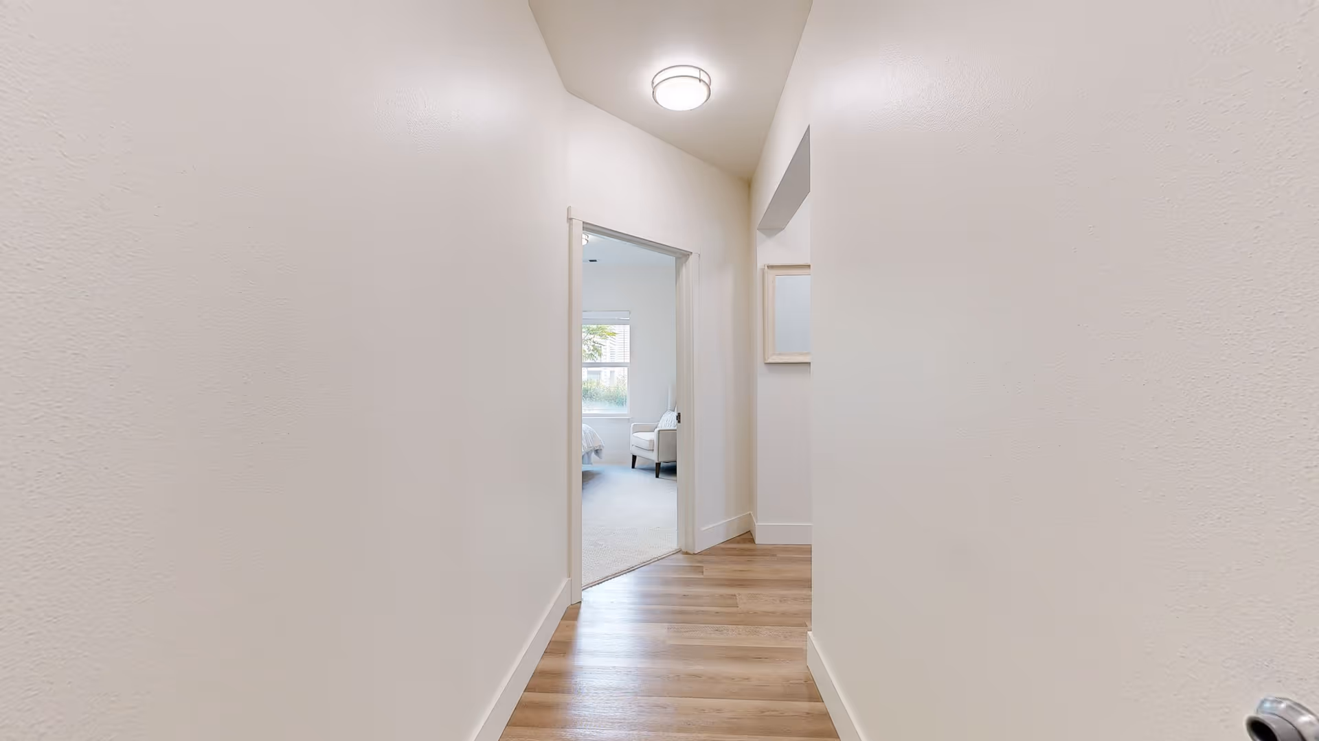 A narrow hallway with light-colored walls and wooden flooring leading to a bedroom. The bedroom has a window with blinds, a bed, and a chair visible. A ceiling light fixture is mounted in the hallway.