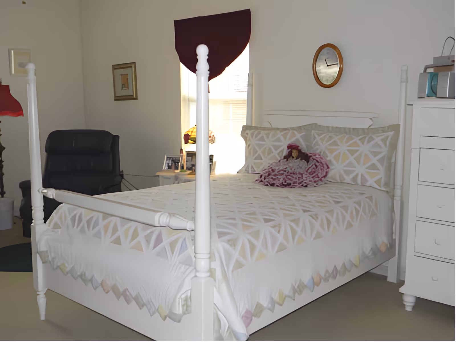 A bedroom with a white four-poster bed featuring a geometric patterned bedspread and pillows. A doll dressed in pink sits on the bed. There is a black recliner chair to the left, a small side table with framed photos and a lamp near the window, and a white dresser with a microwave on top to the right. A round clock hangs on the wall above the bed.