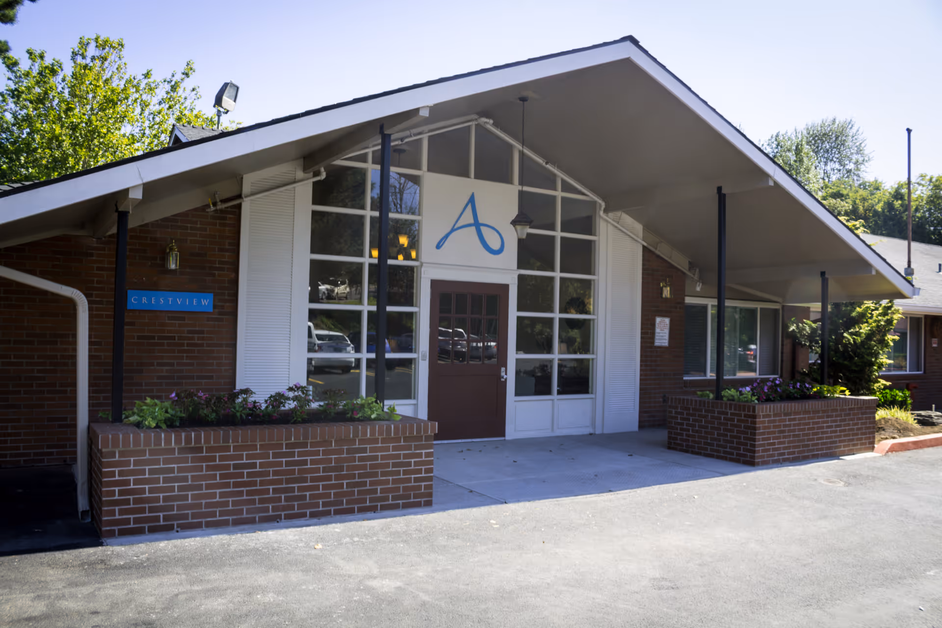 Front entrance of Avamere Crestview of Portland facility featuring a sloped roof, large windows, a brown door, brick planters with flowers, and a blue sign that reads 'Crestview'.