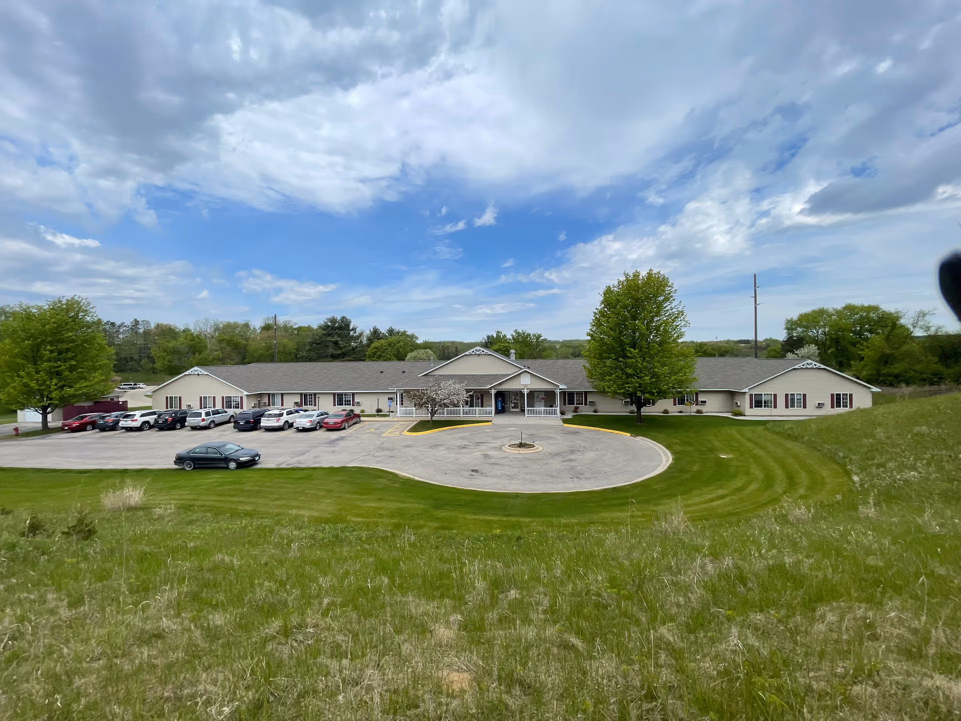 Single-story beige assisted living building with a circular driveway, parked cars and a grassy foreground under a partly cloudy sky.