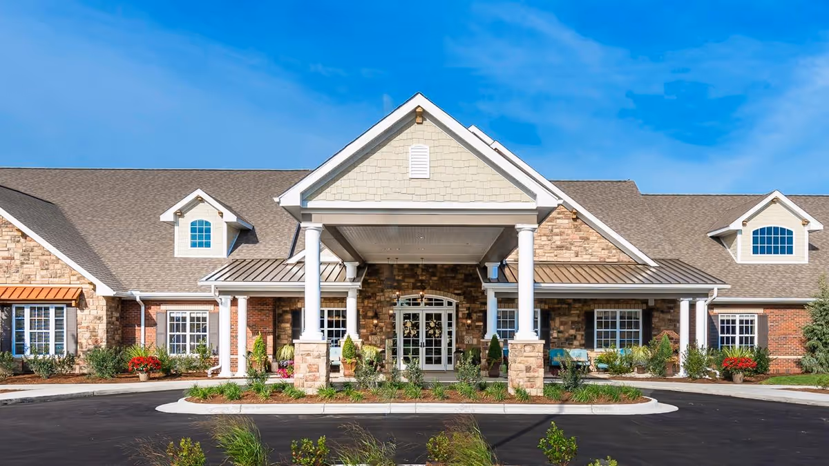 Front exterior view of a senior living facility building with a peaked roof, stone and brick facade, white columns supporting a covered entrance, and landscaped greenery under a clear blue sky.