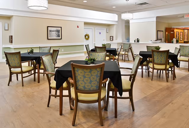 A bright and spacious dining area with multiple square tables covered with black tablecloths, each surrounded by wooden chairs with patterned green and yellow cushions. The room has light-colored walls, wooden flooring, and ceiling lights, with some framed artwork and decorative wreaths on the walls.