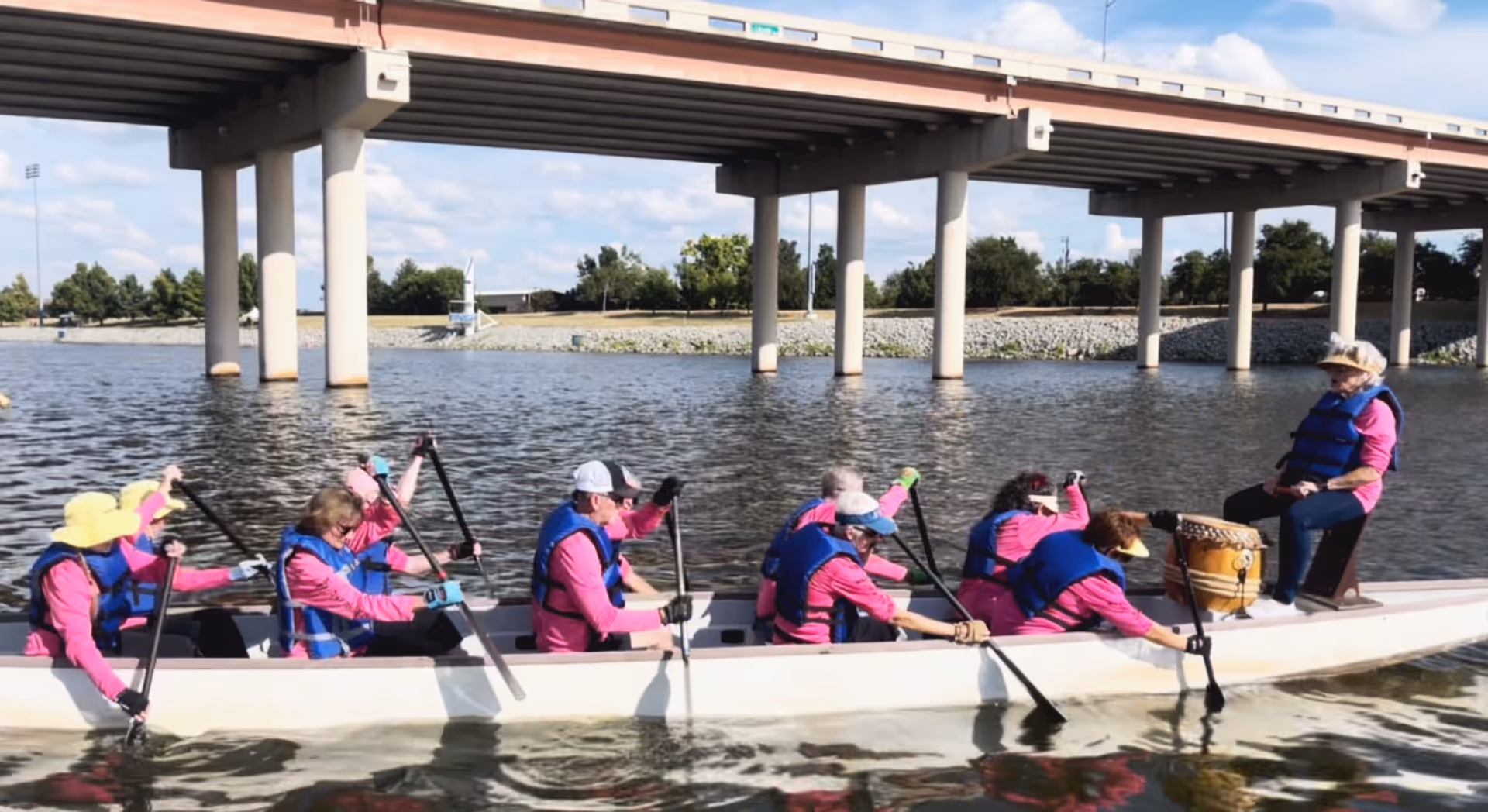 A group of paddlers wearing pink shirts and blue life jackets rowing a long boat on the water beneath a highway overpass.