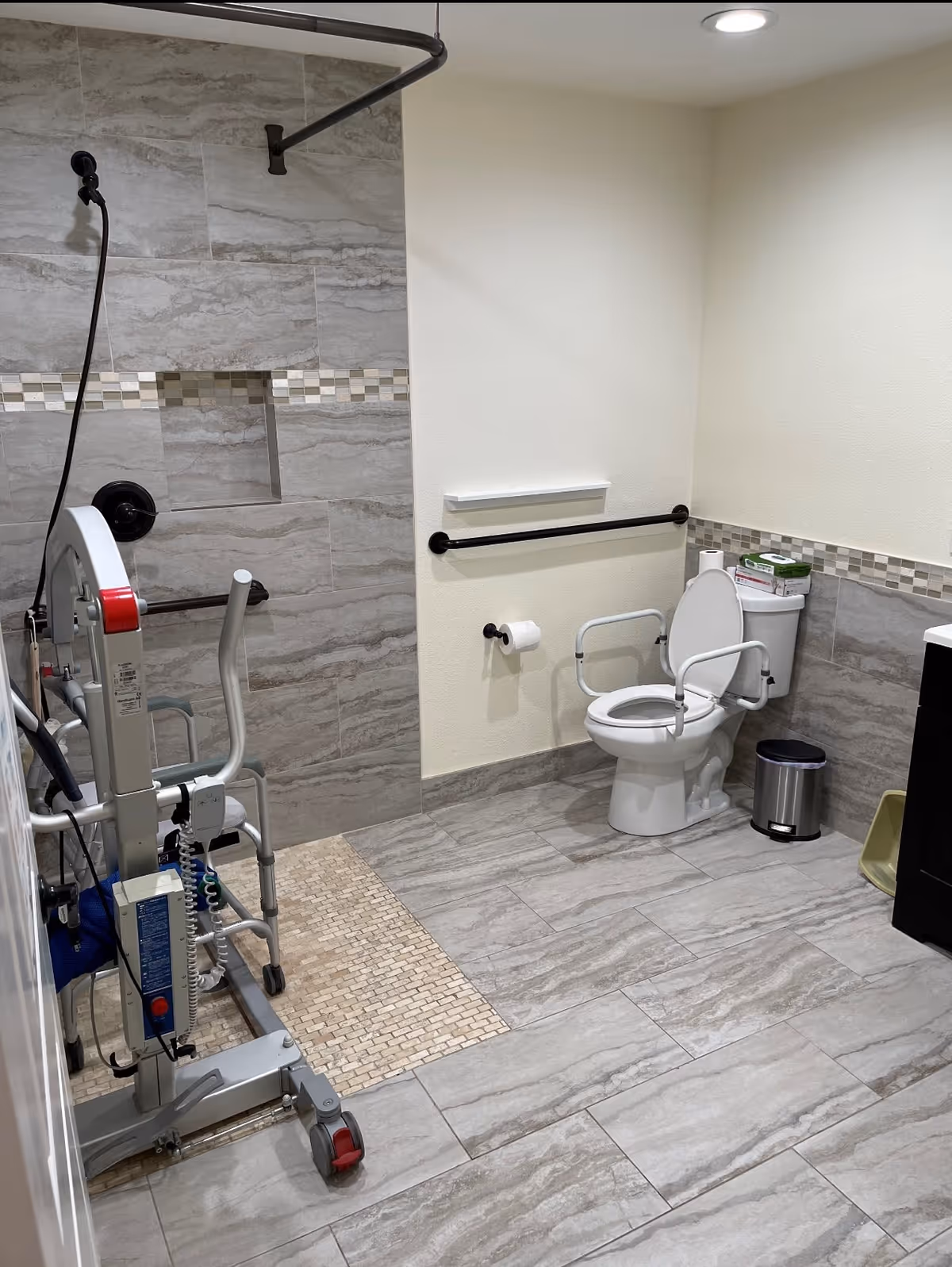 A bathroom designed for accessibility with a toilet equipped with safety rails, a wall-mounted grab bar, and a roll-in shower area with a handheld showerhead. The floor and walls are tiled in neutral tones, and there is a small trash can next to the toilet.