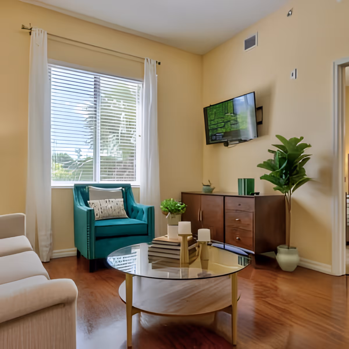 A cozy living room with a beige sofa on the left, a teal armchair with a decorative pillow near a window with white curtains, a round glass coffee table with books and candles, a wooden cabinet with decorative items, a wall-mounted flat-screen TV, and a potted plant in the corner.
