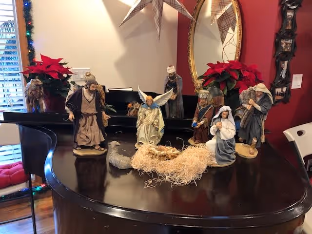 A nativity scene displayed on a dark wooden piano with figurines including Mary, Joseph, baby Jesus in a manger, an angel, the three wise men, and a shepherd. Two red poinsettia plants are placed behind the nativity set, with a mirror and a red wall in the background. Part of a chair and window blinds are visible on the sides.