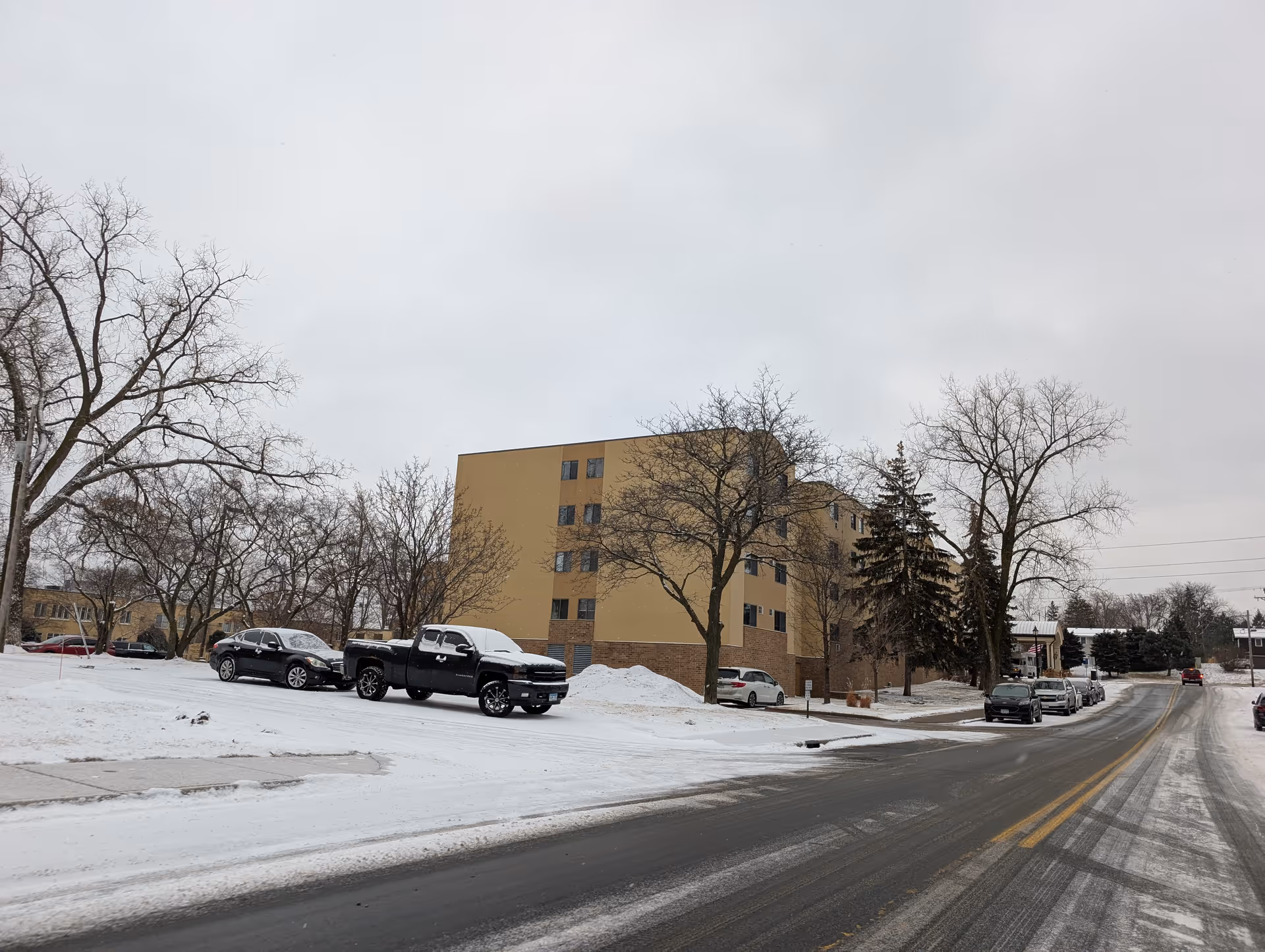 Front exterior of the multi-story beige Chandler Place building in winter with snow on the ground, parked cars, and leafless trees.