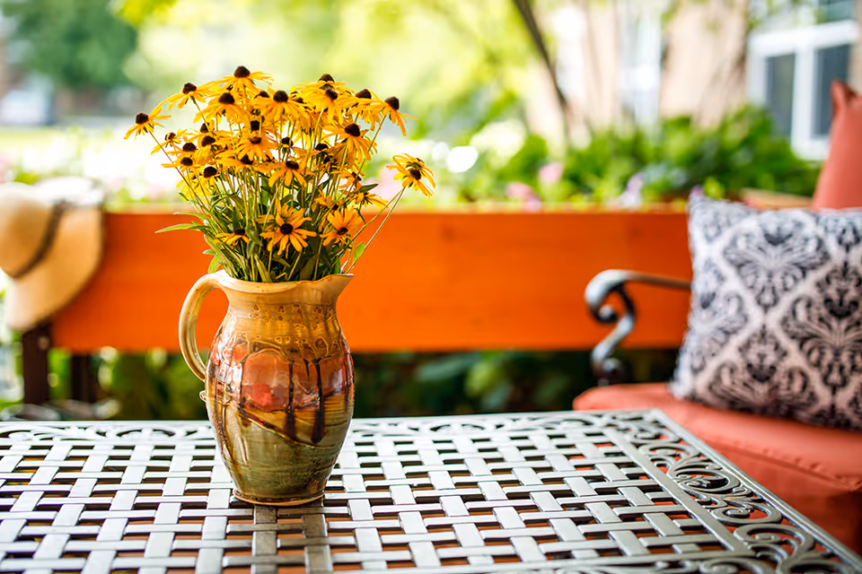 A ceramic pitcher filled with yellow flowers placed on a decorative metal table outdoors, with a cushioned chair and greenery in the background.