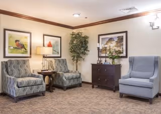 A cozy seating area with upholstered armchairs, a side table, framed artwork, a potted plant, and a small chest in a lounge.