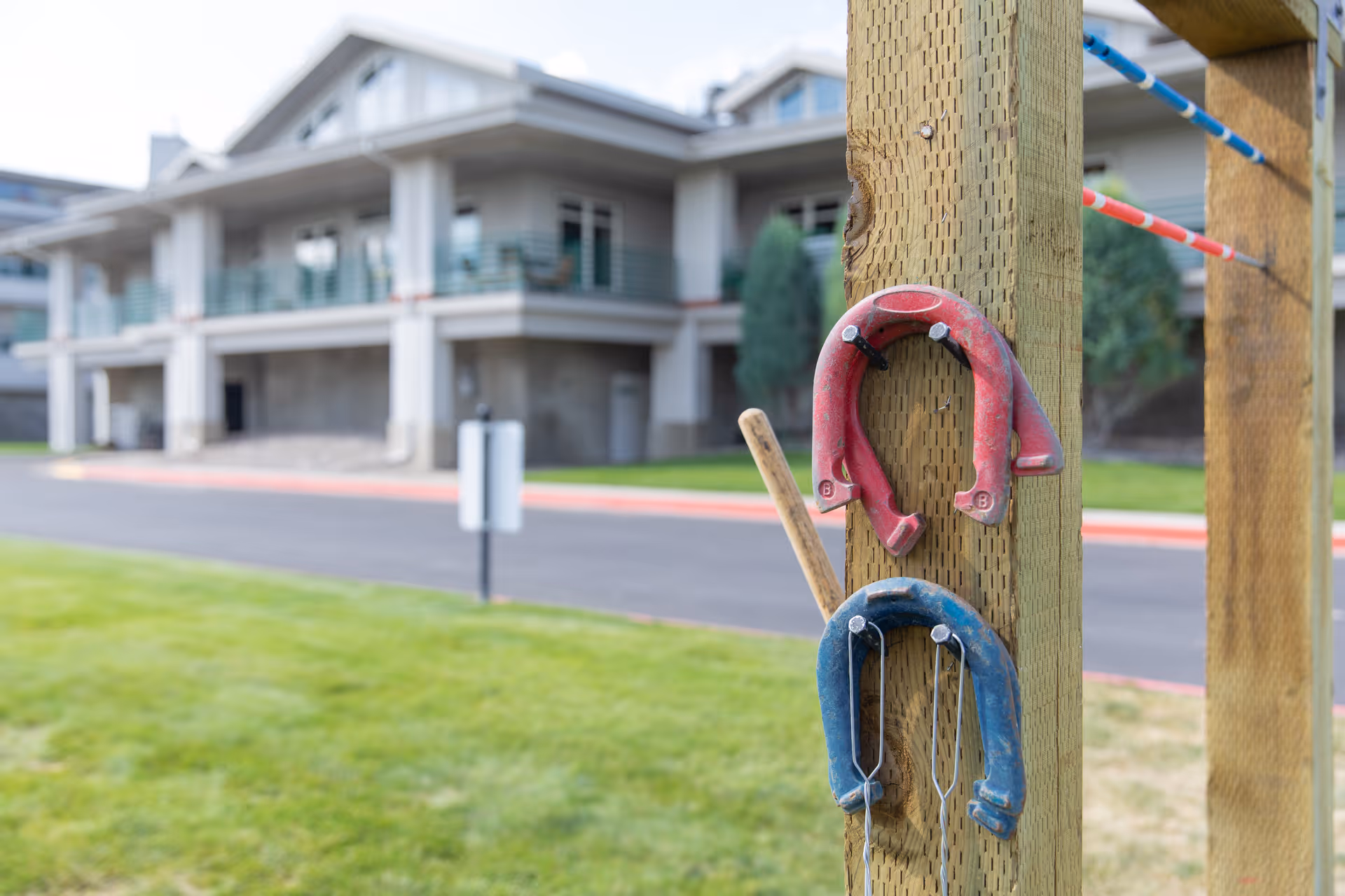 Close-up of a wooden post with red and blue horseshoes and a wooden stick hanging on it, with a senior living facility building and green lawn in the background.