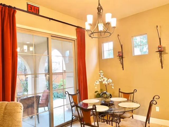 A dining area with a round glass table set with four white placemats and surrounded by four metal chairs with cushions. There is a decorative plant centerpiece on the table. The room has beige walls with two small square windows and two wall-mounted candle holders. A chandelier with five lights hangs above the table. To the left, there is a sliding glass door with red curtains leading to an outdoor patio with chairs and greenery. An exit sign is mounted above the door.