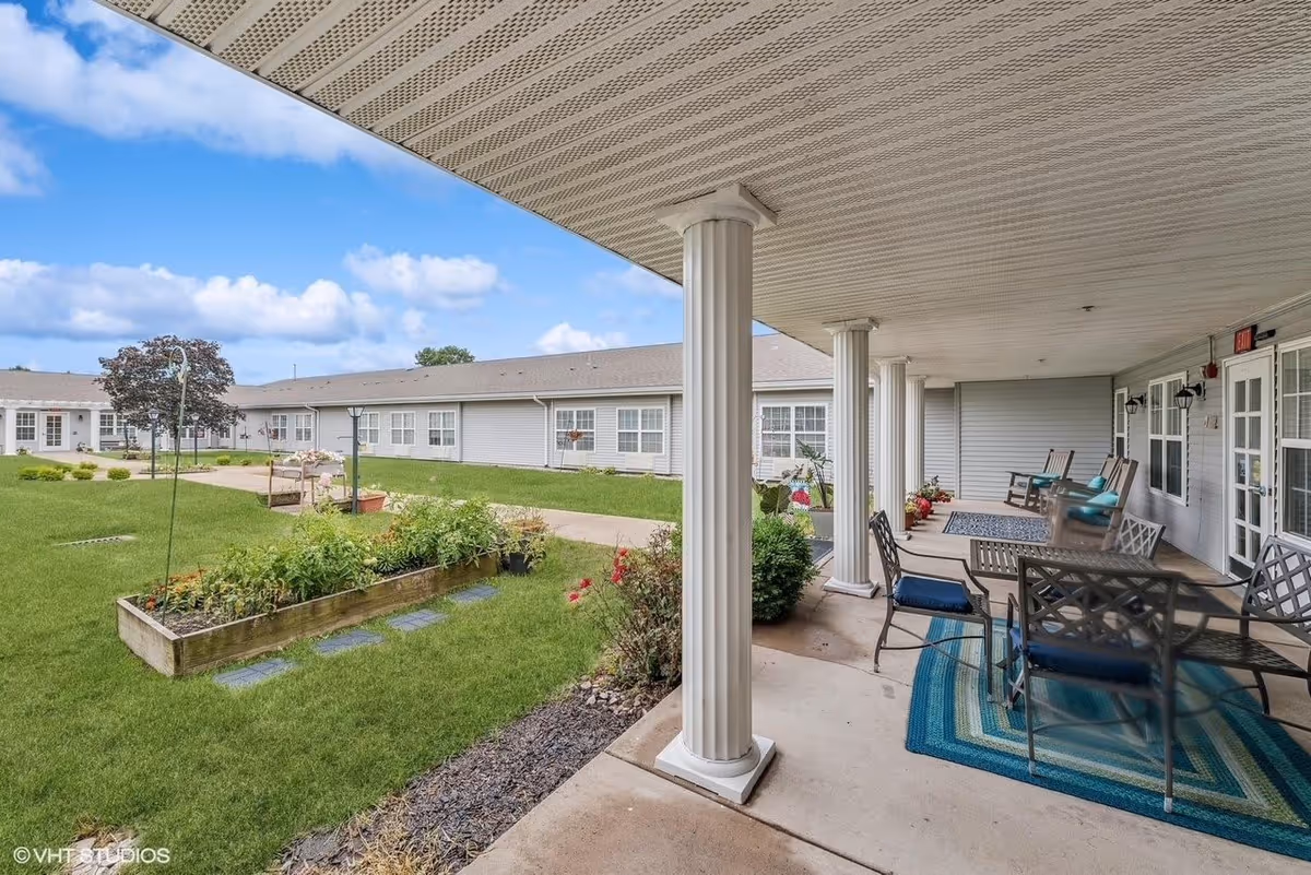 Covered outdoor patio area with white columns, metal chairs and tables on blue rugs, overlooking a green lawn with garden beds and a building with multiple windows in the background under a partly cloudy sky.