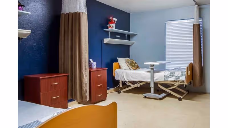 A shared resident bedroom with two adjustable beds, bedside dressers, shelving, privacy curtains, and an overbed table.