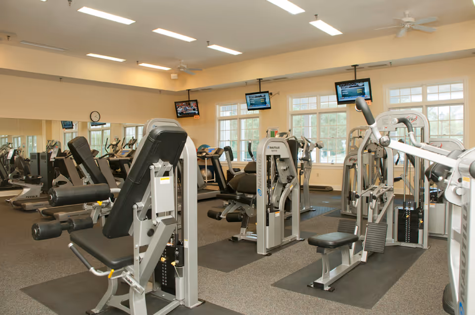 A well-lit fitness room with various exercise machines including weight machines and cardio equipment. The room has large windows allowing natural light to enter, mirrors on one wall, and multiple televisions mounted near the ceiling.