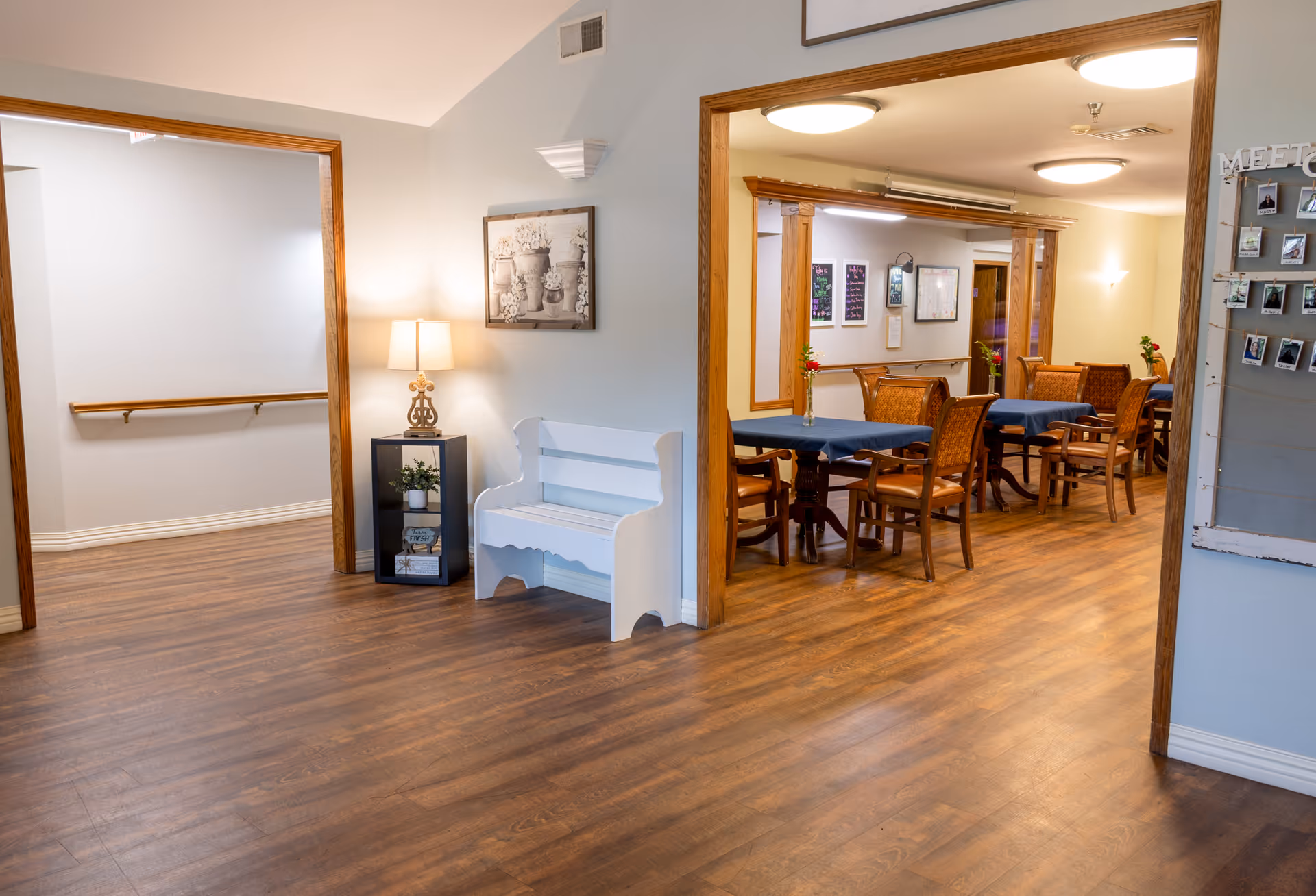 Interior view of a senior living facility showing a hallway with wooden flooring, a white bench, a small black shelf with a lamp and decorative items, and an adjacent dining area with tables covered in blue tablecloths and wooden chairs. The walls are light-colored with framed pictures and a bulletin board with photos.