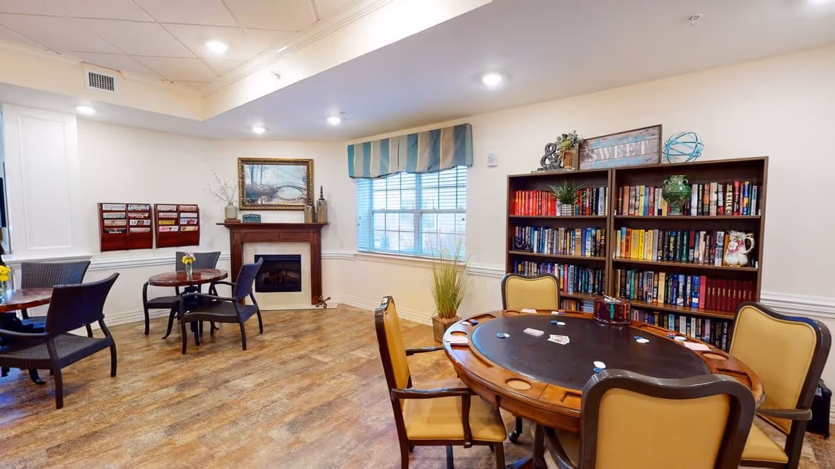 A cozy common room with a round card table surrounded by four chairs, a bookshelf filled with books and decorative items, a window with striped valance, a fireplace with a framed painting above it, and two small round tables with chairs near the wall.