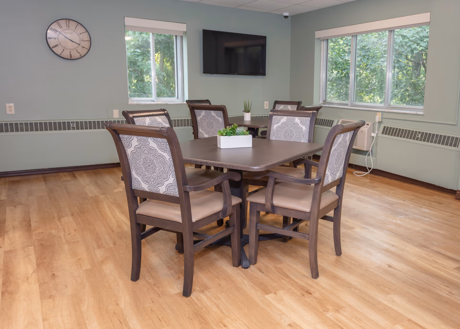 A dining area with a square wooden table surrounded by six cushioned chairs with patterned backs. The room has light green walls, two windows showing greenery outside, a wall clock, a mounted flat-screen TV, and a small potted plant centerpiece on the table. The floor is light wood, and there is an air conditioning unit below one window.