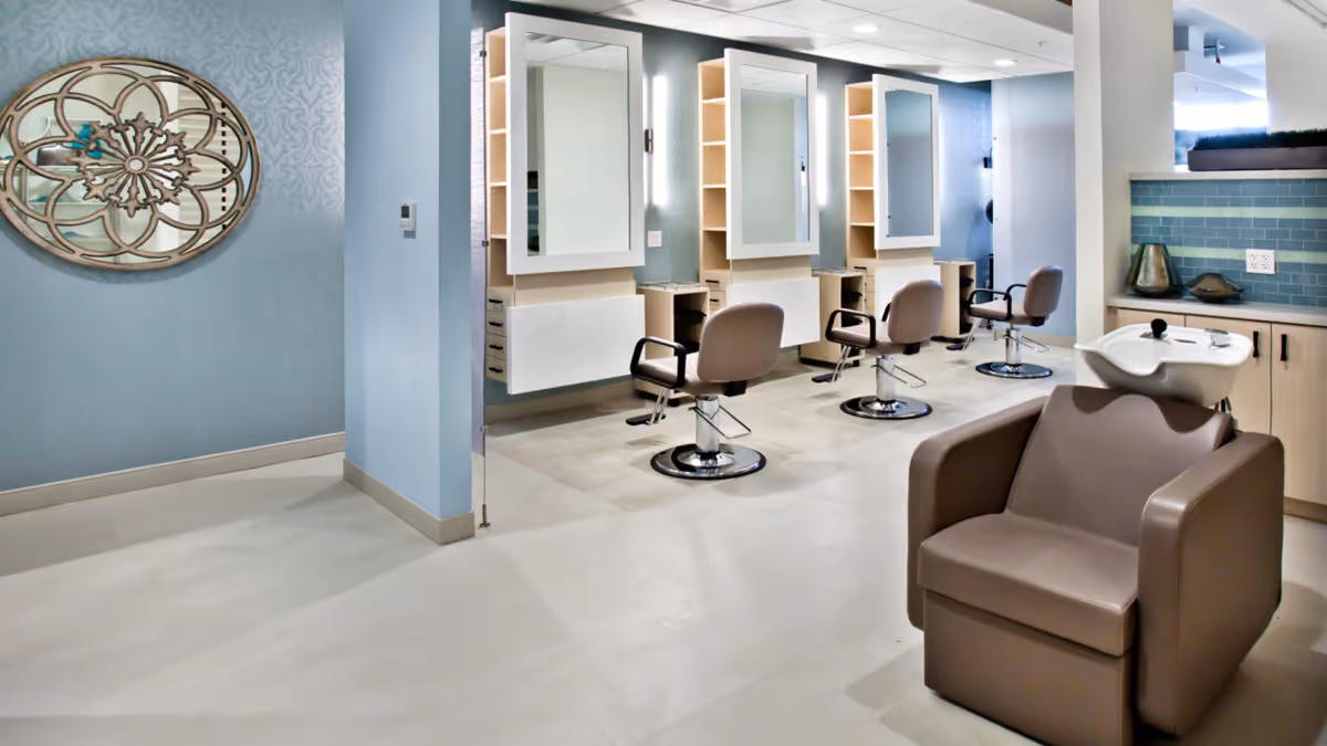 A modern salon area inside a facility with three styling stations, each with a mirror, shelves, and a salon chair. In the foreground, there is a brown shampoo chair with a white sink. The walls are painted light blue with a decorative round wall mirror on the left side.