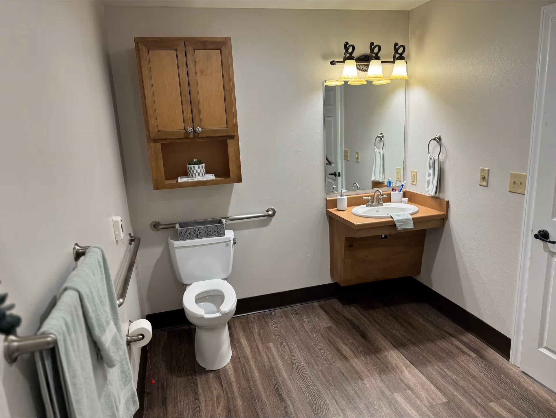 A clean and accessible bathroom with a toilet, a wooden cabinet mounted on the wall above the toilet, a sink with a wooden vanity, a large mirror, and a light fixture with four bulbs above the mirror. There are grab bars near the toilet and a towel hanging on a bar on the left wall. The floor has a wood-like finish.